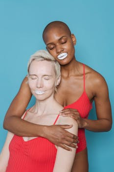 Hairless ethnic lady with closed eyes and white lipstick hugging content fair haired female in red bodysuit in studio